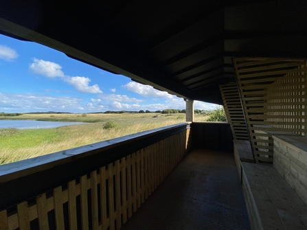 The image shows the interior of a wooden birdwatching hide overlooking a tranquil landscape. A long, low wooden bench runs along one wall, offering a place to sit and observe the scenery. The hide’s large window provides unobstructed views of a calm body of water, grassy wetlands, and a distant horizon under a bright blue sky with fluffy white clouds. The overall atmosphere is peaceful and serene, designed for quiet observation of nature.