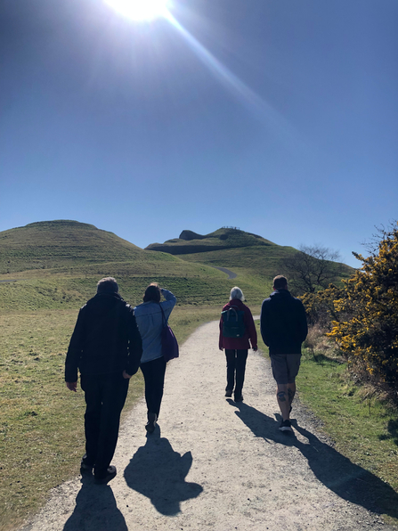 Four people walking away from camera towards Northumberlandia with blue skies above.